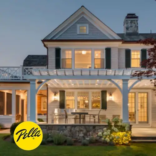 Shingle-style home at dusk with many lit windows and a white pergola, with Pella logo