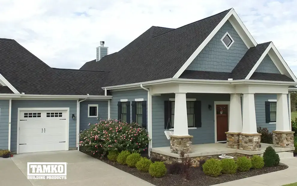 Blue shake-sided home with dark shingle roof, garage and stone-based porch columns