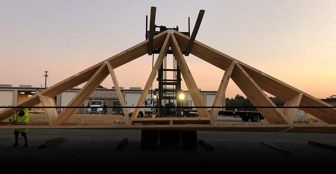 Forklift lifting a large wood roof truss at dusk as a worker looks on
