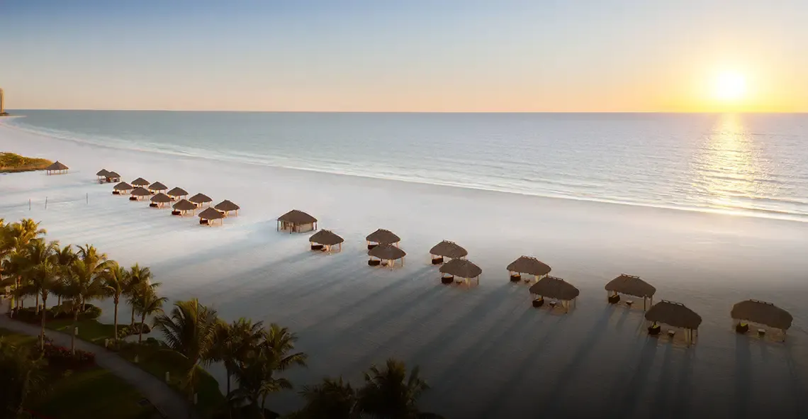 Beach at sunset lined with thatched umbrellas and palm trees