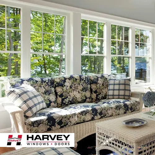 Sunroom with white double-hung windows, wicker sofa and Harvey Windows + Doors logo