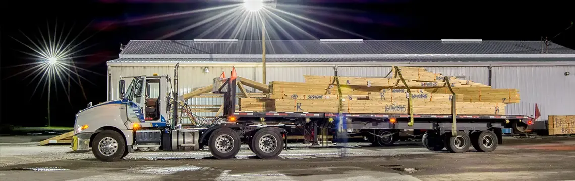 Flatbed truck loaded with bundled trusses outside a plant at night