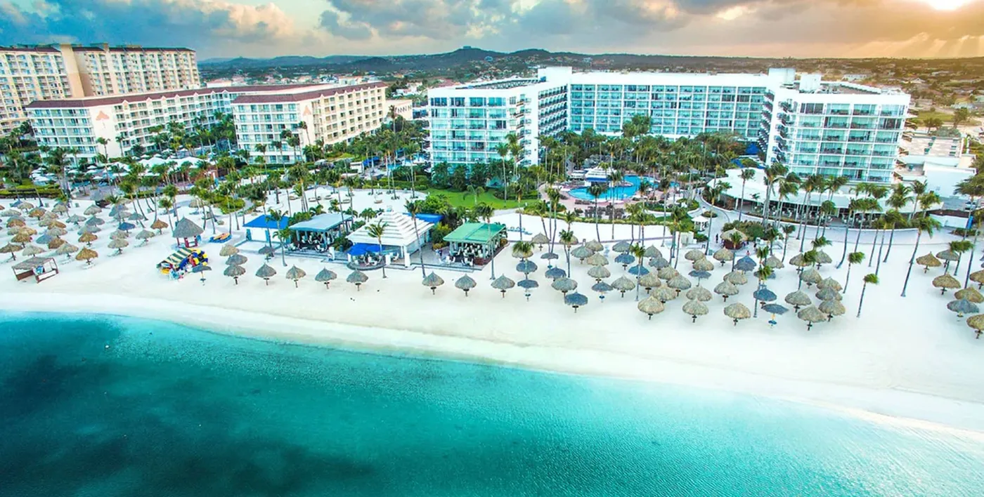 Aerial view of a beachfront resort with palm trees, thatched umbrellas and turquoise water